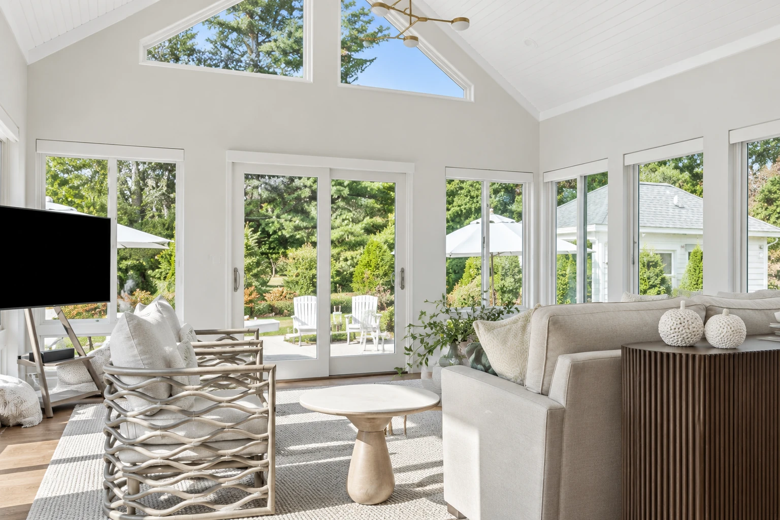 Bright sunroom with vaulted white ceiling, wall of windows, and neutral seating overlooking a green garden.